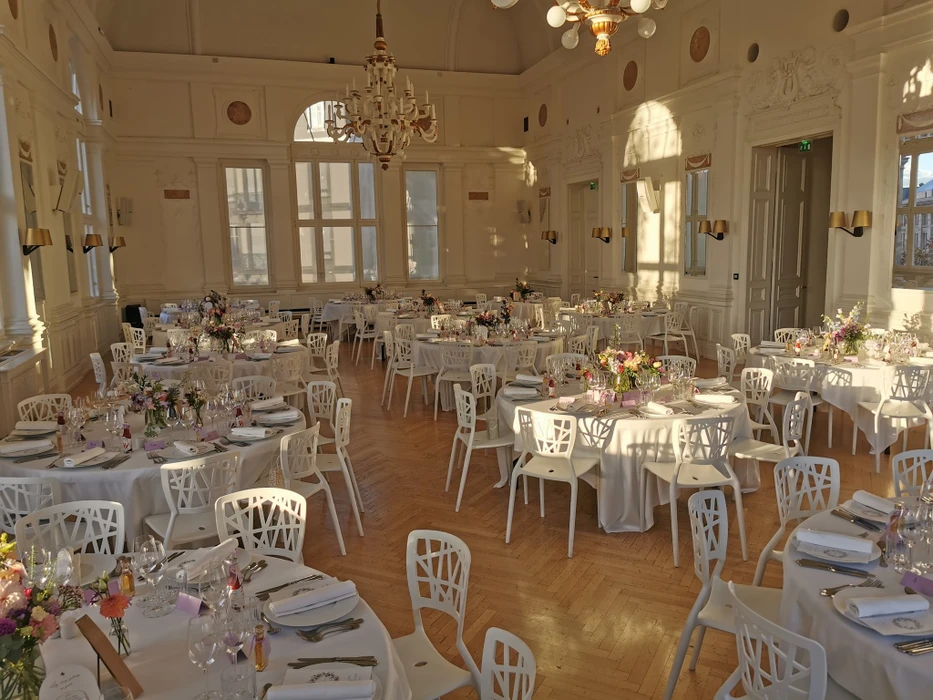 Famille multigénérationnelle célébrant un anniversaire dans cuisine moderne, femme blonde au centre tenant gâteau avec bougies allumées, entourée de six personnes portant chapeaux de fête colorés, sourires et ambiance chaleureuse, décoration festive avec guirlandes